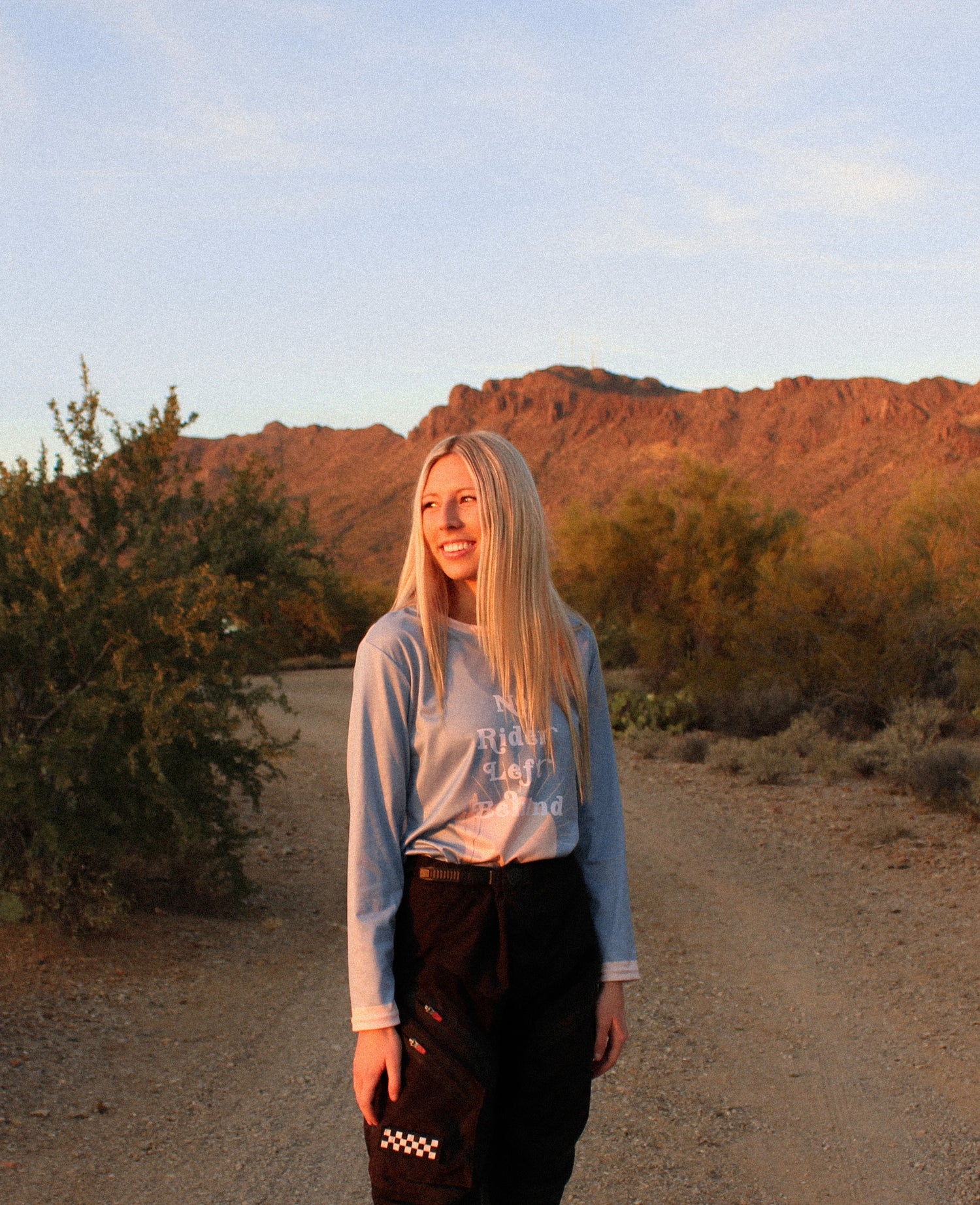 Woman standing in the desert with a light blue jersey on that says "No Rider Left Behind"