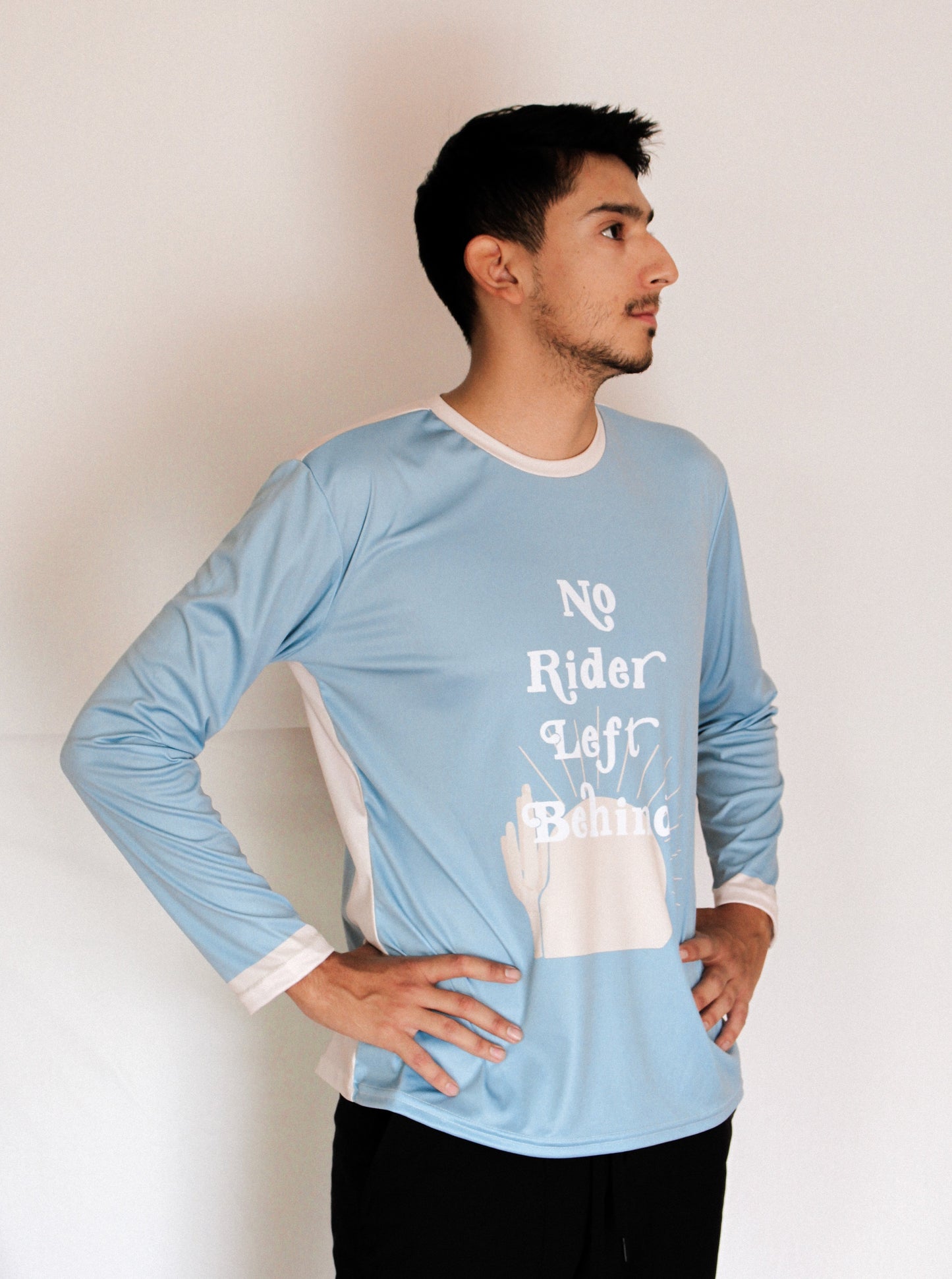 Boy with brown hair standing with his hands on his hips wearing a light blue jersey that says "No Rider Left Behind" with a tan cactus and sun in the background of the jersey