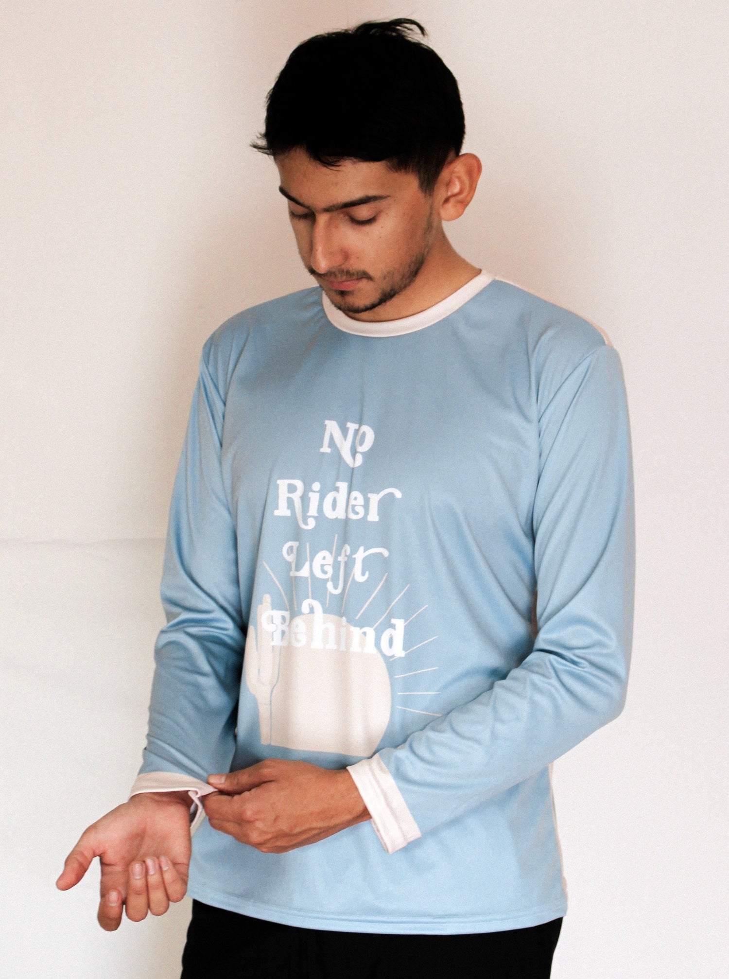 Boy with brown hair standing looking down at his wrists wearing a light blue jersey that says "No Rider Left Behind". There is a tan colored sun and cactus on the jersey.